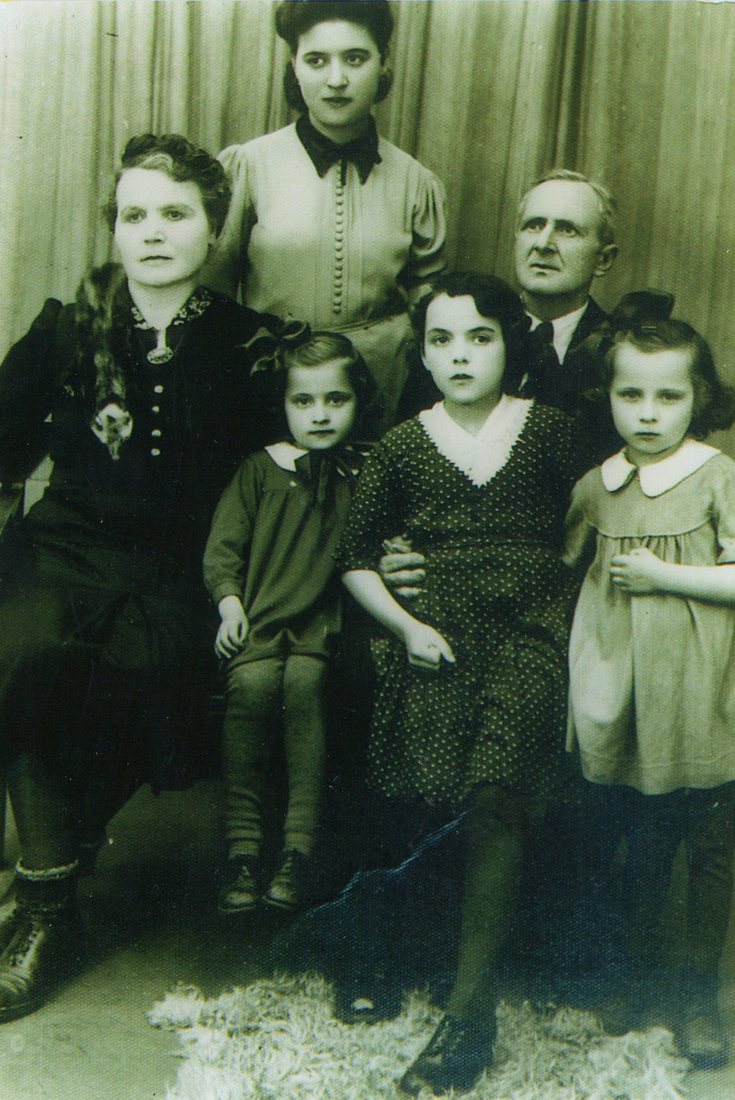 Black-and-white studio photograph of six people in front of a draped curtain: a seated older woman, a young woman standing behind her, an older man, and three young girls in the front.